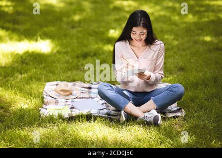 Sorridente ragazza asiatica che prende appunti in blocco note mentre si siede all'aperto nel parco Foto Stock