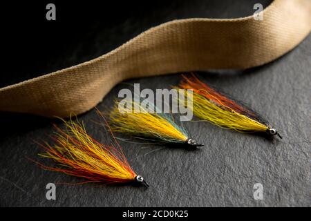 Tre mosche di salmone che erano probabilmente fatte in casa su uno sfondo di ardesia scuro accanto alla cinghia da una vecchia scatola di attacco di vimini. Da una collezione di vinta Foto Stock