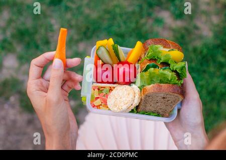 Concetto di cibo sano: Giovane donna che mangia dal pranzo scatola riempito con sandwich, croccanti, frutta e verdura all'aperto; fuoco selettivo, vista dall'alto Foto Stock