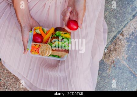 Concetto di cibo sano: Giovane donna che mangia dal pranzo scatola riempito con sandwich, croccanti, frutta e verdura all'aperto; fuoco selettivo Foto Stock