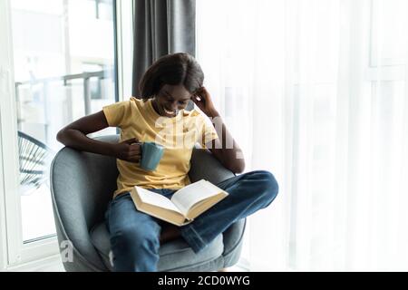 Home tempo libero. Happy black girl leggere libro e bere caffè, seduta in vimini sedia contro finestra in soggiorno, copia spazio Foto Stock