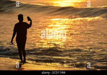 Giovane uomo che prende un auto-ritratto con il suo smartphone sulla riva al tramonto, spiaggia Puri; Puri, Odisha stato, India Foto Stock