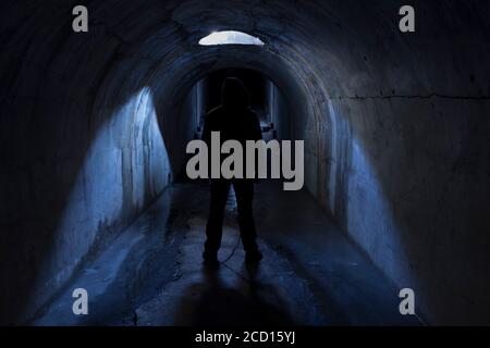 Una silhouette di un uomo con cappuccio in un tunnel sotterraneo scuro vicino ad un portello nel soffitto, illuminato dalla luce. Foto Stock