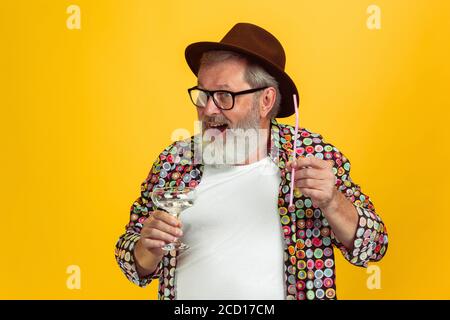 È il momento di bere. Ritratto di uomo hipster senior in occhiali isolati su sfondo giallo studio. Concetto di stile di vita per anziani con tecnologia e gioia. Colori alla moda, per sempre giovani. CopySpace per il tuo annuncio. Foto Stock