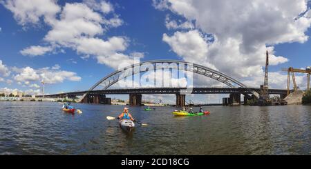 Nuovo ponte ad arco attraverso il fiume Dnieper, in primo piano barche con figure di persone, i volti sono chiusi, Kiev, Ucraina, panorama Foto Stock