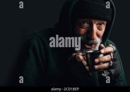 Vecchio senzatetto con barba grigia che si ricopre di verde decrepito indossare tenere una tazza di tè caldo per riscaldare se stesso in una notte fredda Foto Stock