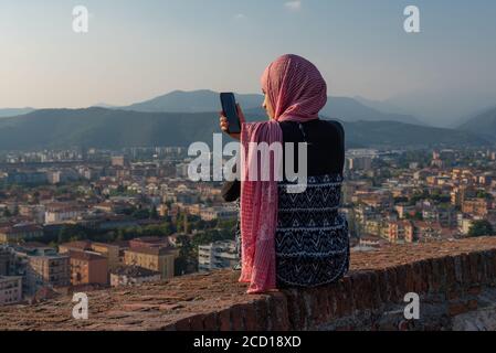 Ragazza egiziana con smartphon sulla terrazza sul tetto con vista aerea della vecchia città italiana Brescia. Foto Stock