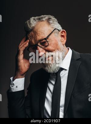 L'uomo d'affari anziano elegante con i capelli grigi pendenti in tuta nera formale, essendo profondo nei pensieri, tiene le palme vicino al mento, isolato sopra il buio Foto Stock