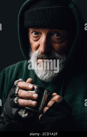 Gli anziani mendicanti di essere congelati all'esterno, bere acqua calda per riscaldarsi, guardando la telecamera isolata su sfondo nero studio. Vecchiaia, senzatetto concep Foto Stock