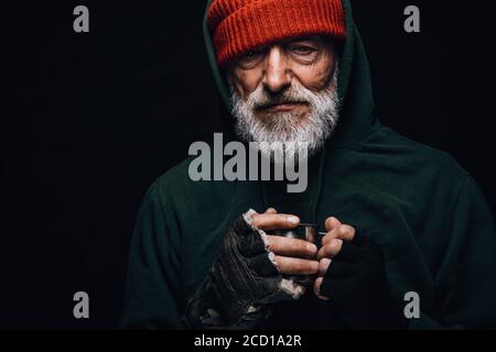 Gli anziani mendicanti di essere congelati all'esterno, bere acqua calda per riscaldarsi, guardando la telecamera isolata su sfondo nero studio. Vecchiaia, senzatetto concep Foto Stock