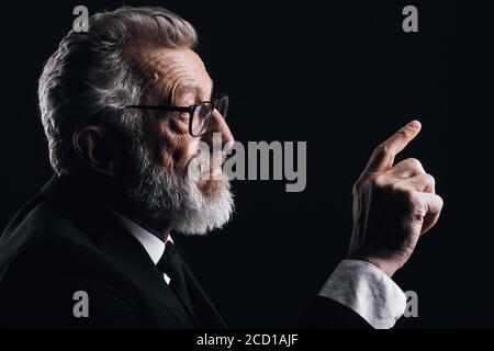 Profilo headshot di intelligente bearded grigio capelli scienziato facendo rapporto in abito nero formale, gesturing con mani isolate su buio Foto Stock