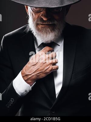 Un vecchio attore maschio che gioca un ruolo di spia inglese, vestito in vestito nero, nascosto dietro il cappello di fedora, avendo un misterioso aspetto elegante. Studio isolato portra Foto Stock