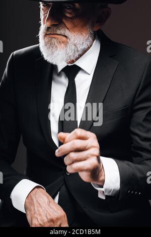 Elegante ritratto per gentiluomo anziano in stile classico con cappello trilby e tuta nera. Moda maschile, look classico concetto. Studio girato al buio Foto Stock