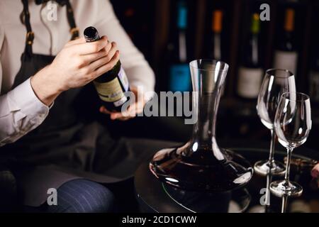 Primo piano di eleganti sommelier in camicia bianca e grembiule che versano vino in decanter. Degustazione di vini. Sommelier composizione della lista dei vini Foto Stock