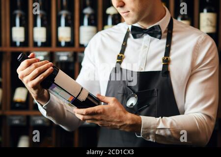 Un piccolo colpo di giovane cavista caucasico vestito di camicia bianca e bowtie lavorando in grande negozio di vite che presenta una bottiglia di vino rosso al cliente Foto Stock