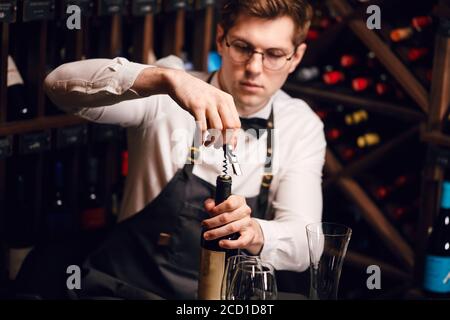 Barista che apre una bottiglia di vino con cavatappi nel ristorante con scaffali di vino. Foto Stock