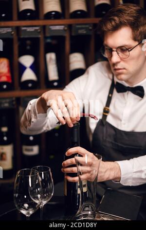 Barista che apre una bottiglia di vino con cavatappi nel ristorante con scaffali di vino. Foto Stock