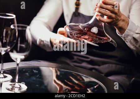 Vista ritagliata del sommelier professionista che tiene in mano il recipiente per mescolare con vino rosso sul tavolo del ristorante. Foto Stock