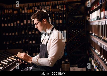 Esperto di vino maschile steward facendo la presa di vino in negozio, scrivendo in taccuino alcune note in camera oscura con file di rack Foto Stock