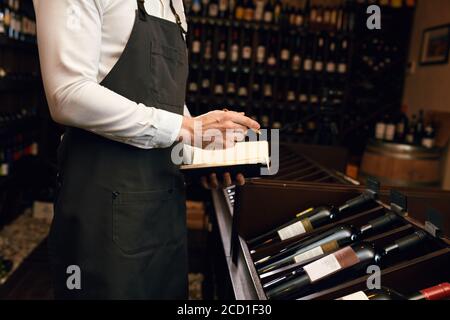 Cavista maschile professionista esamina le bottiglie con vino e fare note al suo blocco note circa le loro caratteristiche e origine in negozio di cantine dove la gente ca Foto Stock