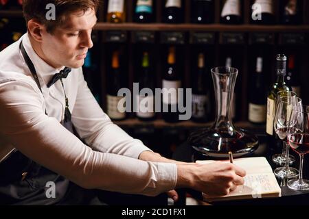 Esperienza di degustazione di vini in hotel o ristorante. Ragazzo professionista o sommelier che tiene un bicchiere di delizioso vino rosso che apprecita la sua tinta con bottiglie co Foto Stock