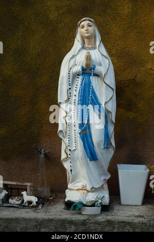 Statua della Vergine Maria simbolo di amore e gentilezza con offerte in piedi su una sporgenza di pietra contro un marrone parete Foto Stock