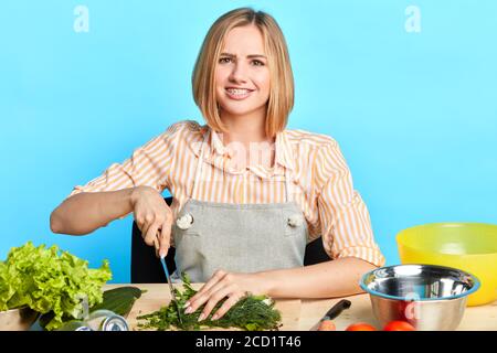 Studio shot di sorridente cuoca con grembiule e coltello di tenuta mentre facendo deliziosa cena per i suoi ospiti, tagliando verdure per insalata, cercando Foto Stock