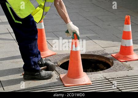 Operaio sopra il portello aperto della fogna su una strada vicino ai coni di traffico. Concetto di riparazione delle acque reflue, delle utenze sotterranee, del sistema di approvvigionamento idrico Foto Stock