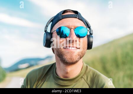 Un uomo fantastico con berretto da baseball, cuffie wireless e occhiali da sole blu che sorridono e ascoltano la musica preferita. Foto Stock