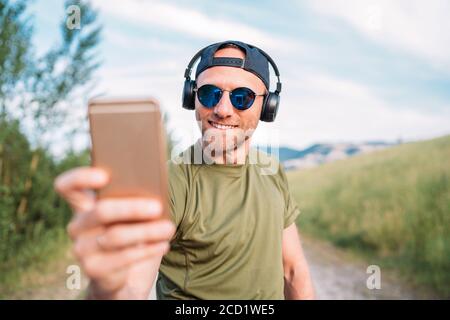 Un uomo fantastico con berretto da baseball, cuffie wireless e occhiali da sole blu che navigano nel suo dispositivo smartphone con playlist per lettore mp3 Foto Stock