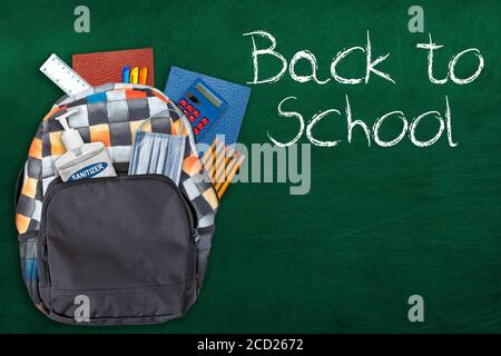 Torna alla scuola concetto di istruzione nella nuova normalità durante la pandemia Covid-19 con zaino ripieno di cancelleria, disinfettante per le mani e maschera viso su cha Foto Stock