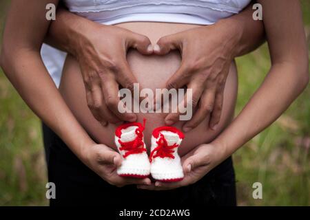 una donna incinta e suo marito abbracciano il loro futuro bambino modellare le mani come un cuore e tenere scarpe per bambini Foto Stock