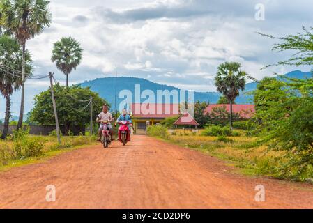 Cambogia paesaggio rurale con due uomini che detengono canne da pesca guidare le moto lungo la strada sterrata con palme di palmyra (Fiabellifero Borassus) sul lato Foto Stock