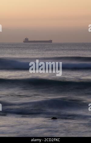 Silhouette di nave da carico all'orizzonte all'alba, onde infrangenti, Durban, Sudafrica, paesaggio, mare, creativo, artistico, trasporto, sfocatura movimento Foto Stock