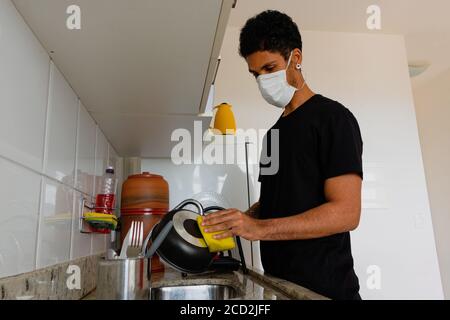 Uomo adulto nero indossando maschera di coronavirus lavando la dishe in cucina. Foto Stock