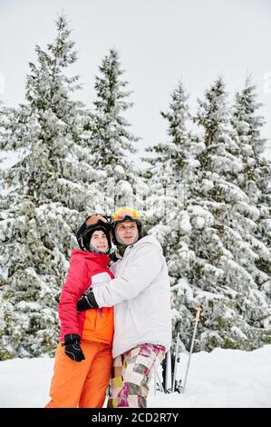 Affascinante uomo e donna in abiti da sci e caschi abbracciano e guardando da parte mentre si trova in piedi nella foresta invernale con alberi di conifere nevosi sullo sfondo. Uomo sciatore abbracciare bella giovane donna e sorridente. Foto Stock