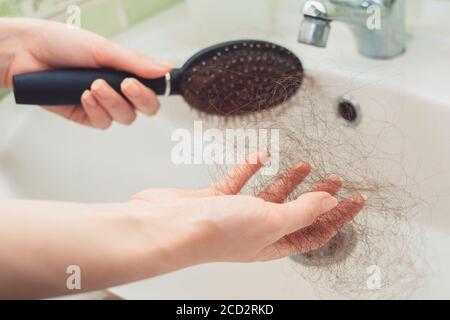 Le mani delle femmine puliscono un pettine sopra il lavandino da un mazzo di capelli strappati. Primo piano delle mani. Il concetto di calvizie e cura dei capelli. Foto Stock