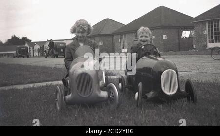 Anni '50, storico, sull'erba fuori degli chalet in mattoni a un piano in un campo di vacanza a Hayling Island in Hampshire, Inghilterra, un bambino e una ragazza che si divertono seduti in pedalò giocattolo 'ride-on'. Foto Stock