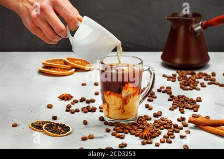 Crema per la preparazione della mano maschile da una tazza bianca in vetro trasparente con caffè freddo, caffè freddo su sfondo grigio e nero, spazio copia. Foto Stock