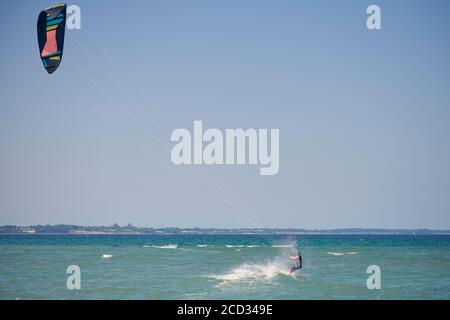 Bruna donna kitesurf o kite boarding che si allontana dal spiaggia di sabbia che fa per acque più profonde in un'estate soleggiata giorno in una vista posteriore del venuto Foto Stock