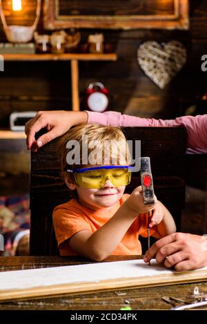 Bambino piccolo che ripara in officina. Professione futura. Capretto con martello e chiodi contro muro di legno. Foto Stock