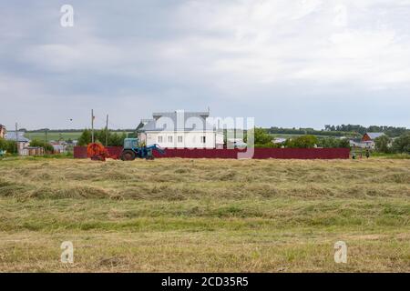 Villaggio di Rybushkino, Russia - 1 luglio 2020: Raccolta di fieno in un villaggio russo. Il trattore blu raccoglie il fieno in grandi bobine rotonde. Villaggio rur Foto Stock