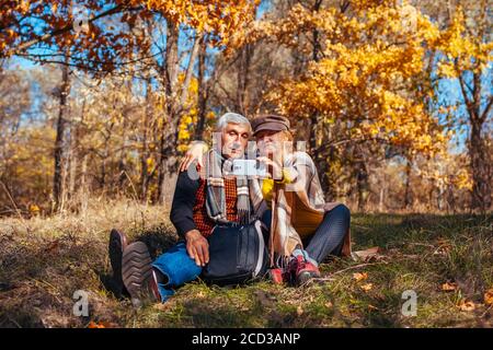 Coppia senior che prende selfie utilizzando smartphone nel parco autunnale. Uomo e donna in pensione godendo della natura e abbracciando Foto Stock