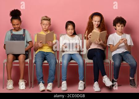 Concetto di e-learning. Ha messo a fuoco i bambini diversi con i gadget e i materiali di studio seduti sulle sedie sopra lo sfondo rosa Foto Stock