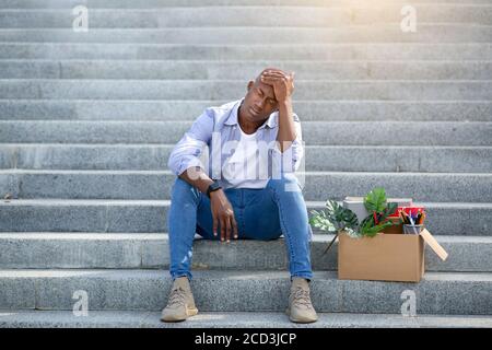 Ragazzo nero depresso con oggetti personali seduti sulle scale dopo aver perso il lavoro, all'aperto Foto Stock