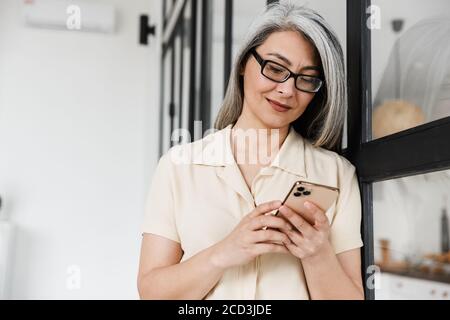 Foto di felice donna asiatica con capelli grigi che indossa occhiali con cellulare telefono in soggiorno Foto Stock