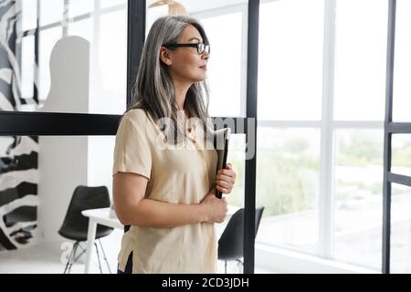 Immagine di una splendida donna di affari matura in piedi vicino alla finestra interna a casa mentre tiene un computer portatile. Foto Stock