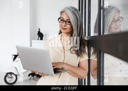 Foto di una donna asiatica con capelli grigi che indossa occhiali che lavora con un computer portatile in soggiorno Foto Stock