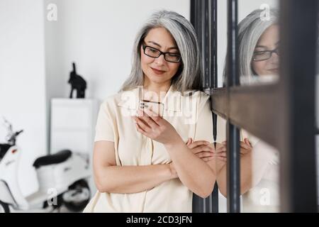 Foto di felice donna asiatica con capelli grigi che indossa occhiali con cellulare telefono in soggiorno Foto Stock