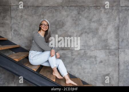 Foto di felice donna asiatica dai capelli grigi che indossa occhiali sorridenti mentre seduto su scala a casa Foto Stock